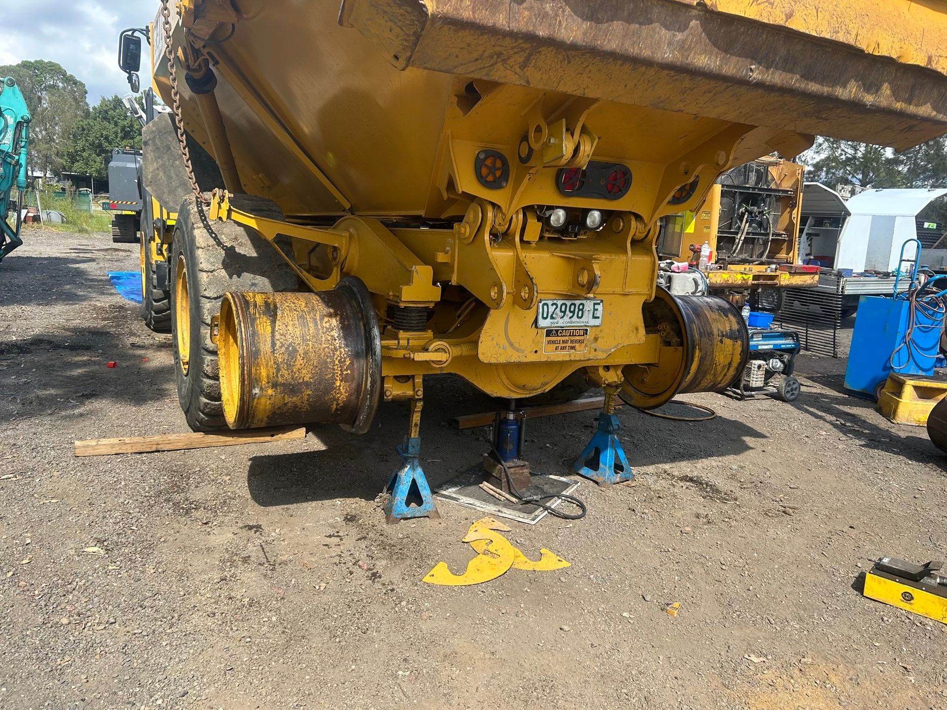 Yellow dump truck lifted on blue jack stands in a gravel yard, with tools and equipment nearby.