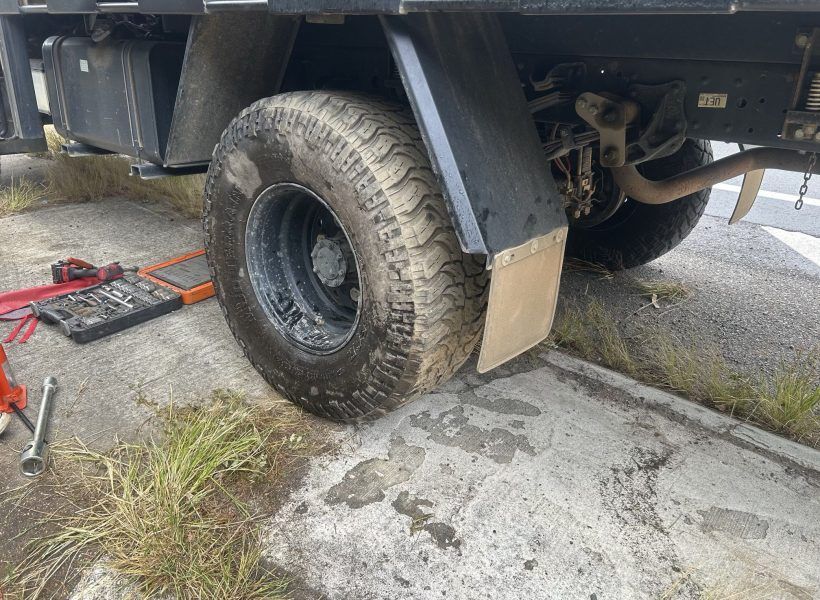 Truck wheel on a curb with a jack and tools nearby, beside a wet driveway and grass.