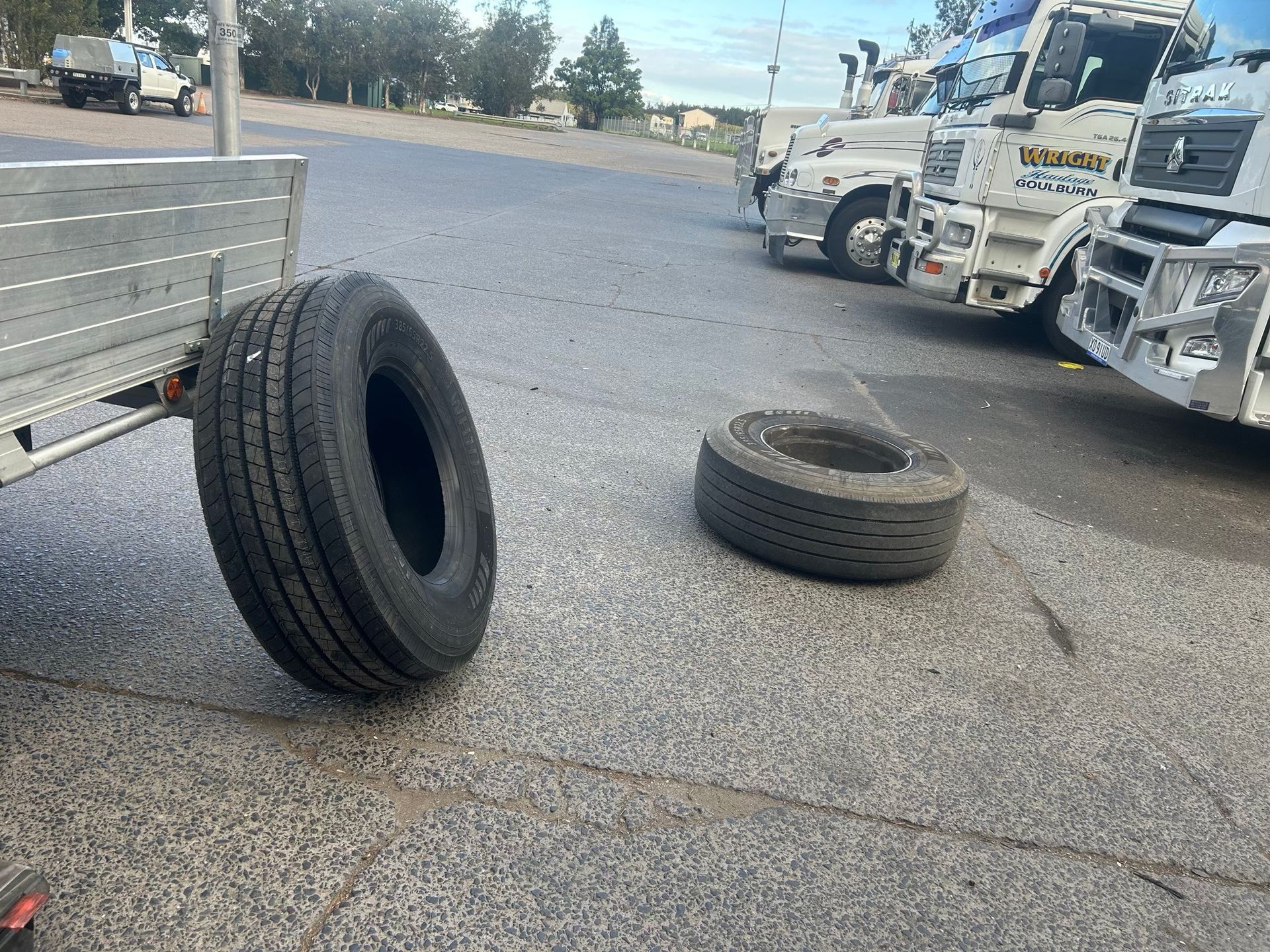 Two truck tires lying on a roadside near parked heavy trucks and a trailer in a gravel lot