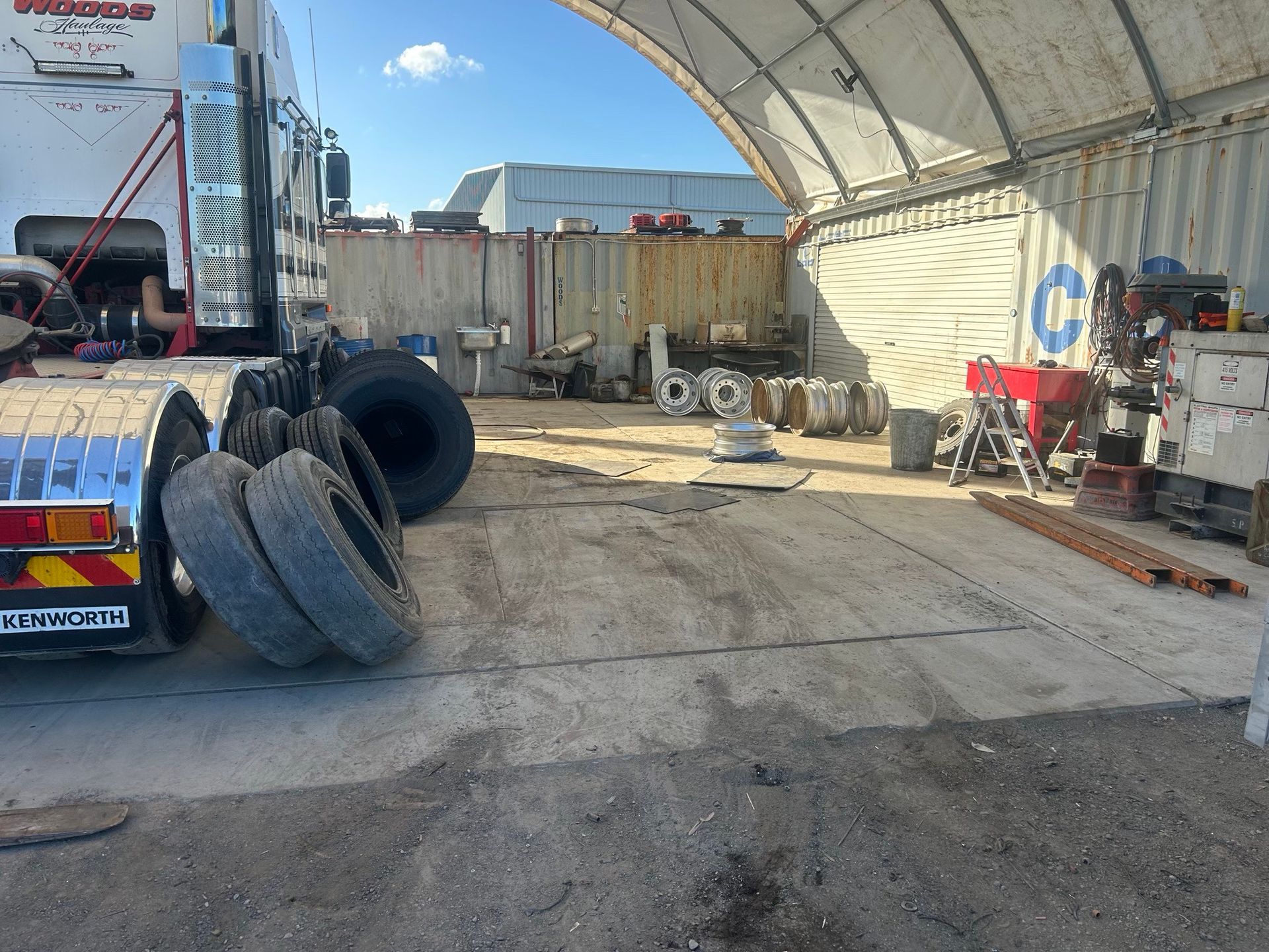 Outdoor industrial yard with stacked tires, shipping containers, and a large white canopy under a blue sky