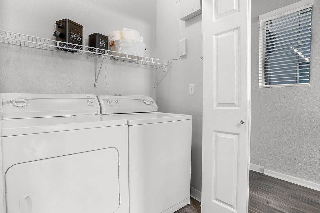 White top-loading washer and dryer side-by-side in a laundry room with a wire shelf above at Solaire Luxury Apartments, offering Albuquerque apartments for rent.