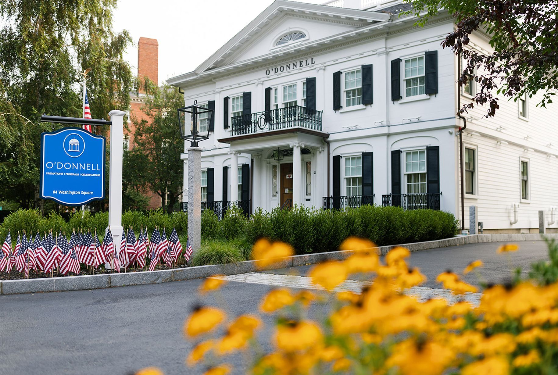 White building with black shutters, sign, and small American flags; yellow flowers in foreground.
