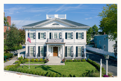 White Greek Revival style building with black shutters, American flag, and manicured lawn.