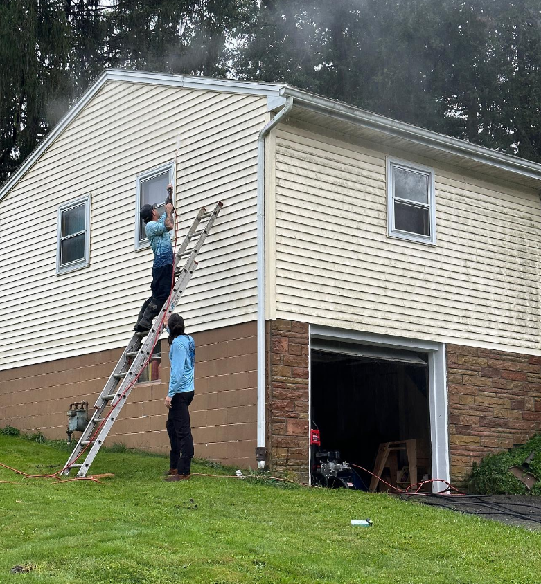 Two people on ladders painting a beige two-story house with a brick lower level and open garage.