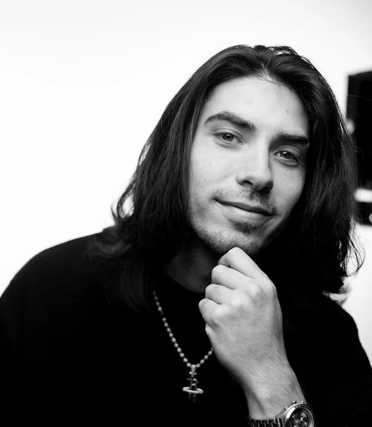 Black-and-white portrait of a person with long hair, resting their chin on a hand and wearing a necklace.