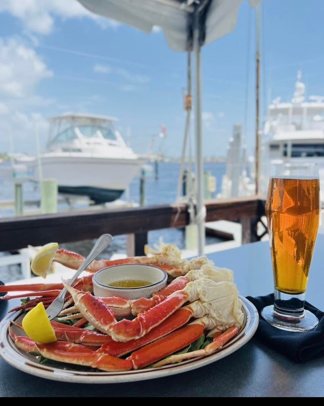 Crab legs and shrimp on a plate with beer, on a waterfront table overlooking boats and blue sky.