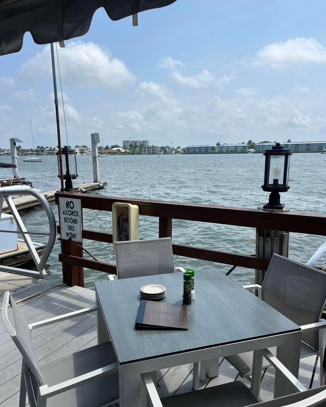 Waterfront patio with white chairs and table overlooking a calm harbor on a sunny day