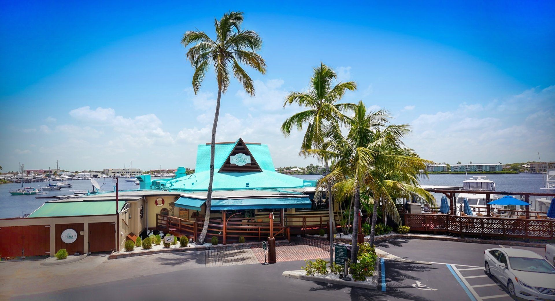 Tropical waterfront restaurant with teal roof, palm trees, and docked boats under a bright blue sky