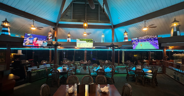 Dimly lit restaurant with a central stage, blue neon accents, and large TVs showing sports.