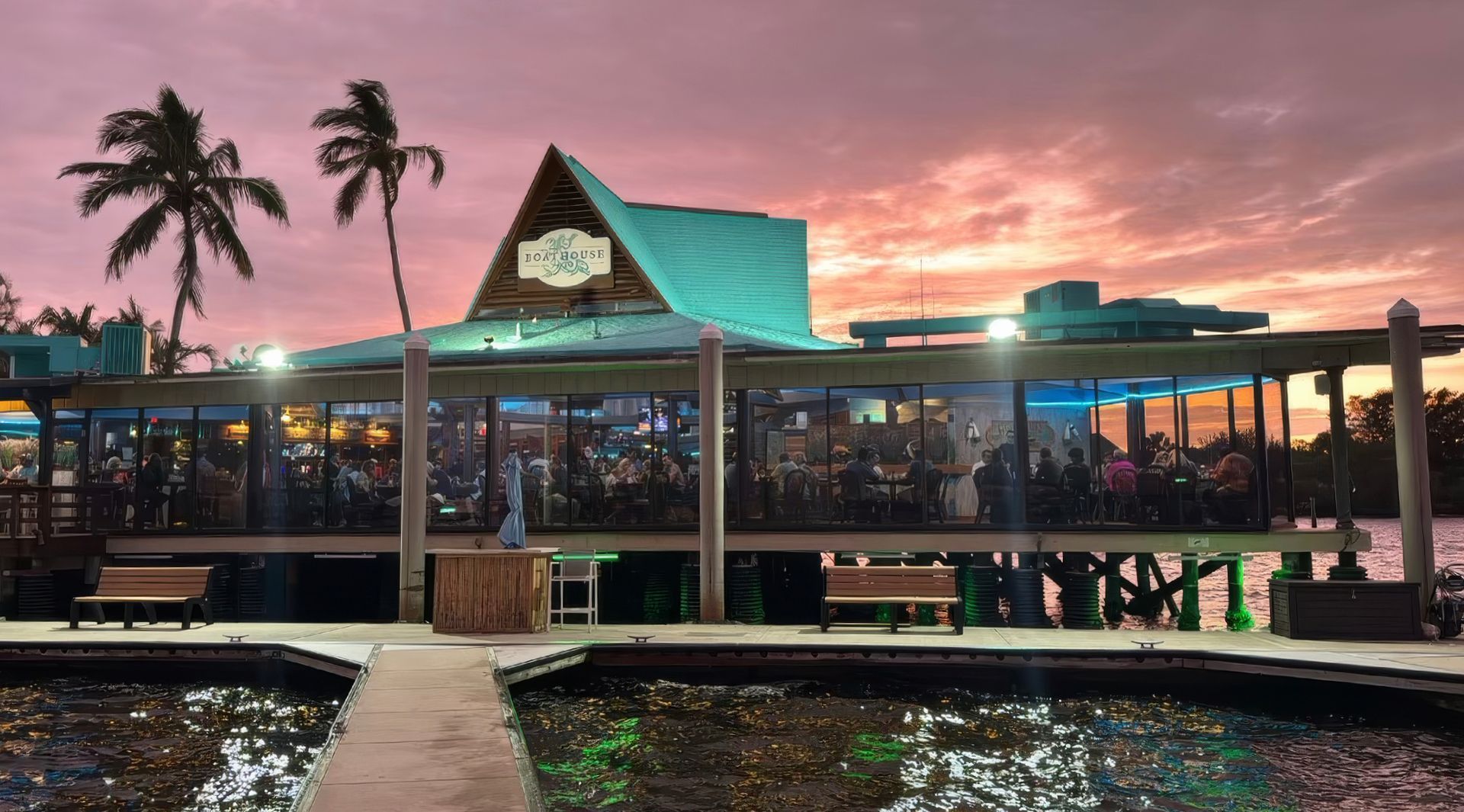 Tropical waterfront restaurant at sunset with pink sky, palm trees, and glowing lights under a teal-roofed pavilion