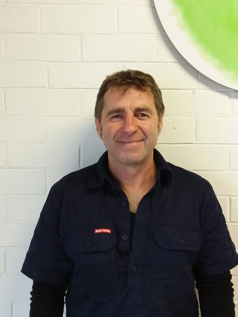 A Man in A Blue Shirt Is Smiling in Front of A White Brick Wall — Sunworks Eco Plumbing In Torquay, VIC