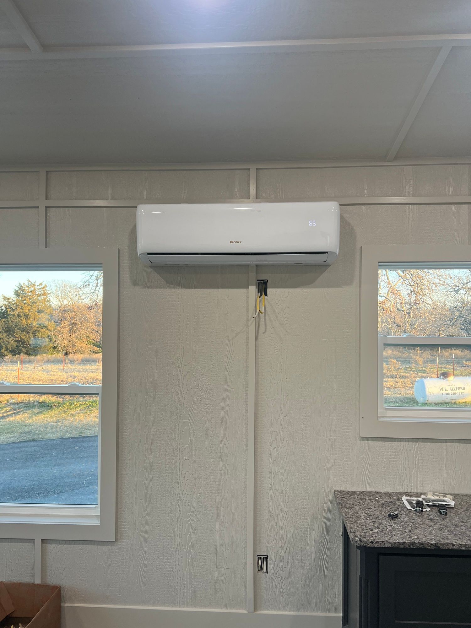 A white air conditioner is hanging from the ceiling in a room with two windows.