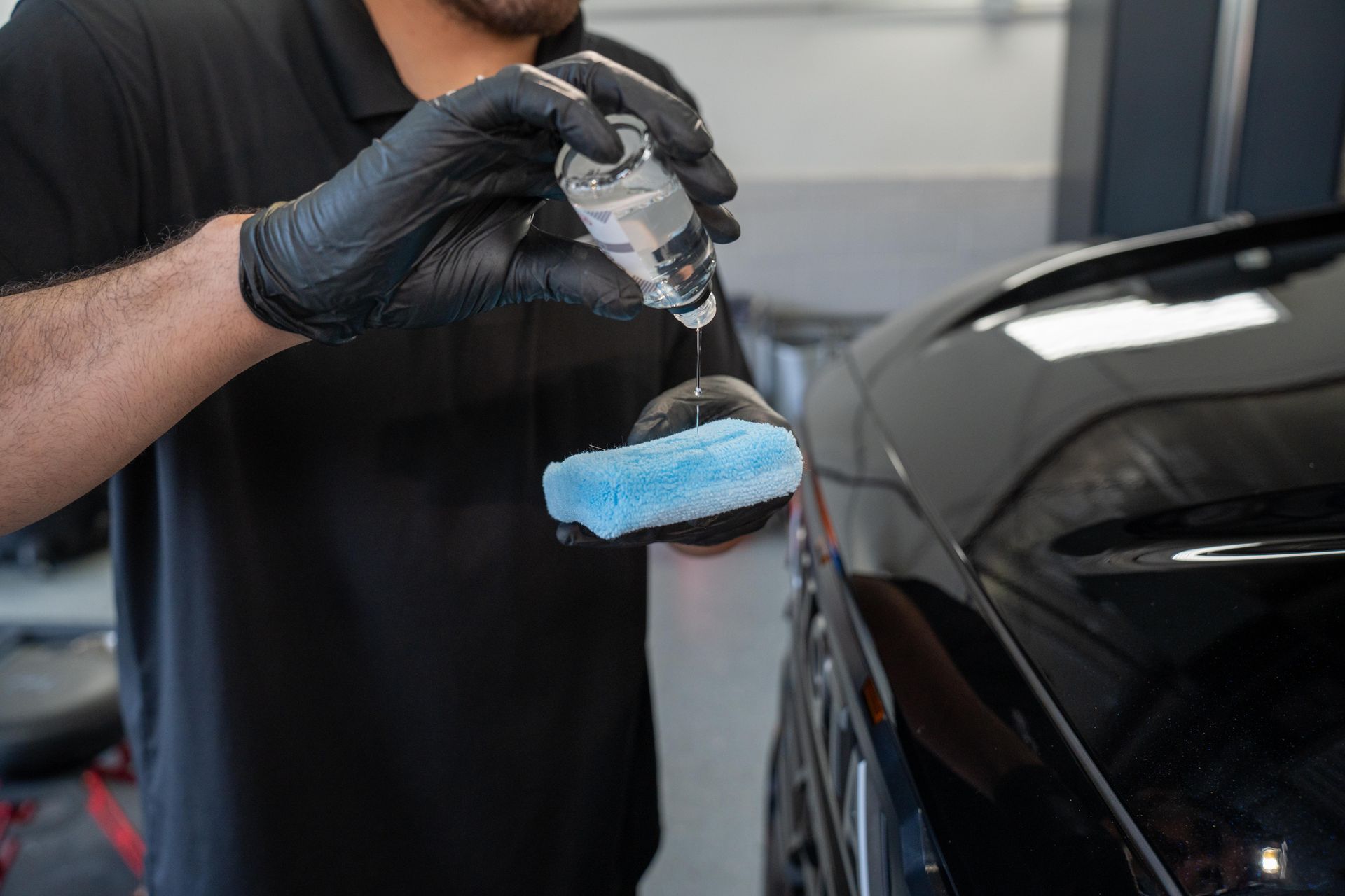 A man wearing black gloves is applying a coating to a black car.
