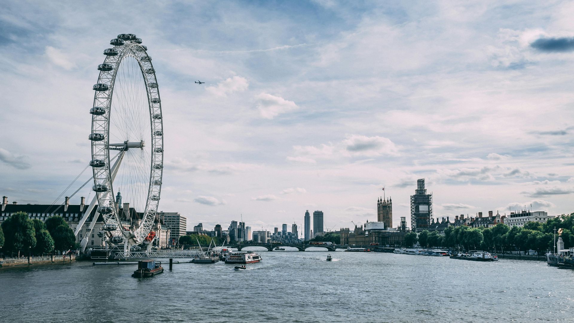 London Eye on the Thames with boats, city skyline, and cloudy sky