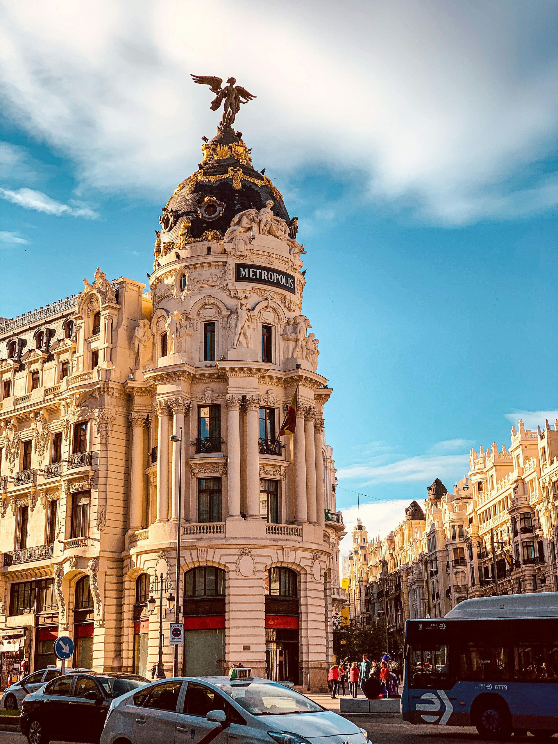 Ornate historic building at a busy city intersection under a blue sky, with traffic and pedestrians nearby