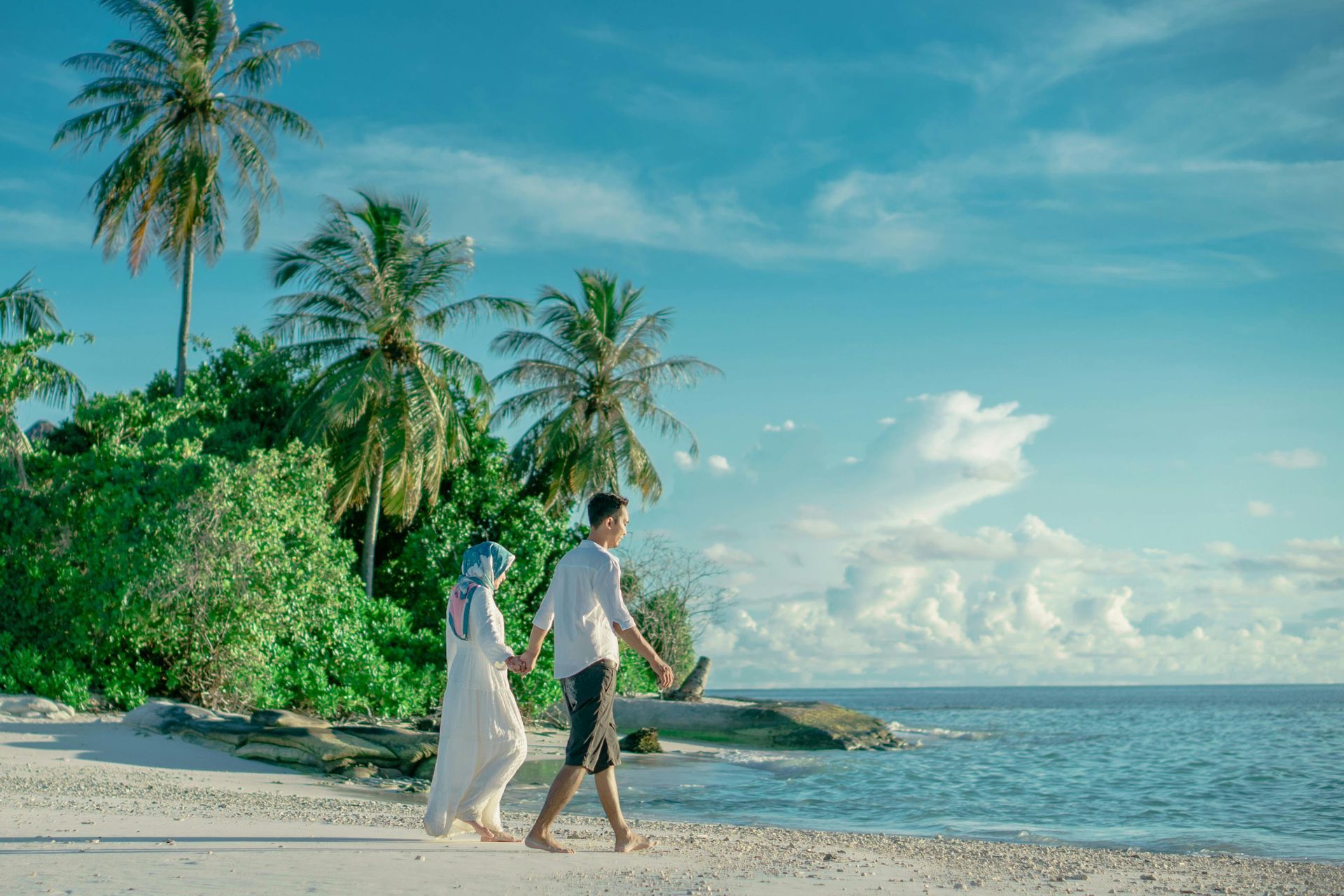 Two people walking along a sandy tropical beach beside clear blue water and palm trees