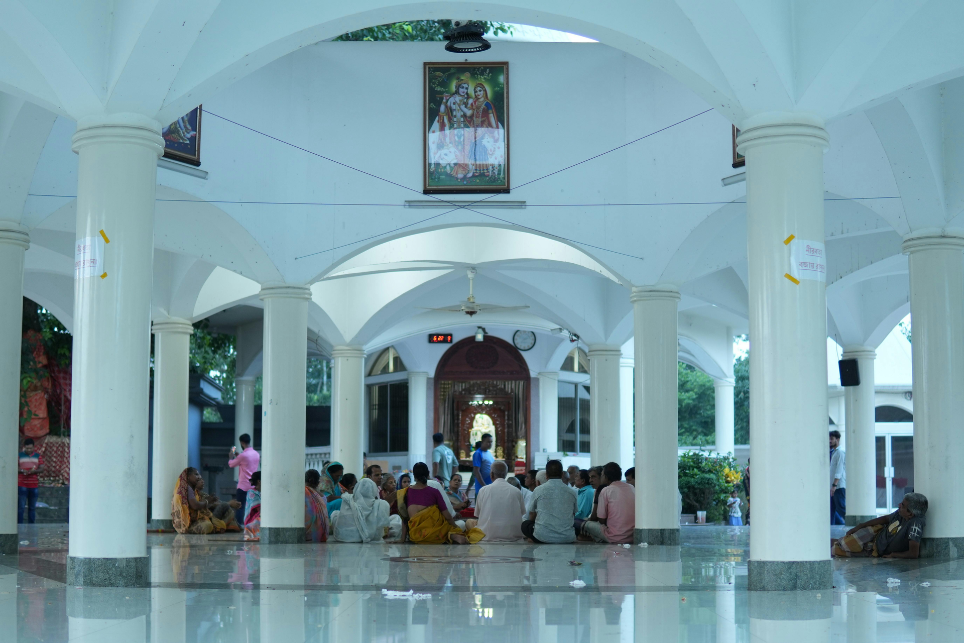 Interior of a white-pillared hall with people seated in prayer or gathering at the far end.