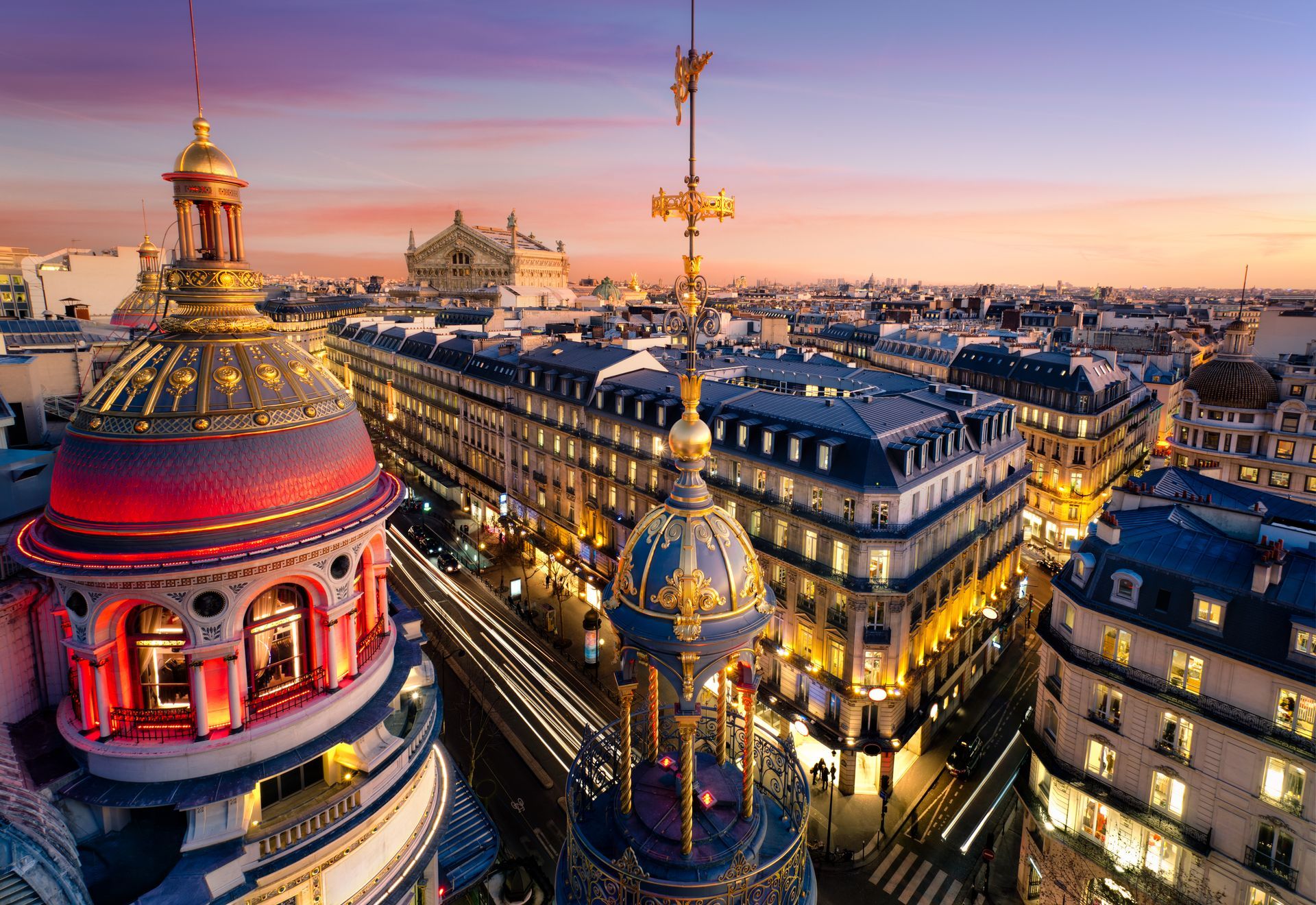 Paris rooftop skyline at dusk with illuminated domes, historic buildings, and streetlights along a city avenue