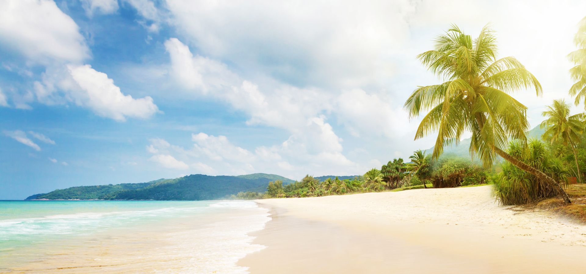 Tropical beach with turquoise water, white sand, palm trees, and distant green hills under a bright sky