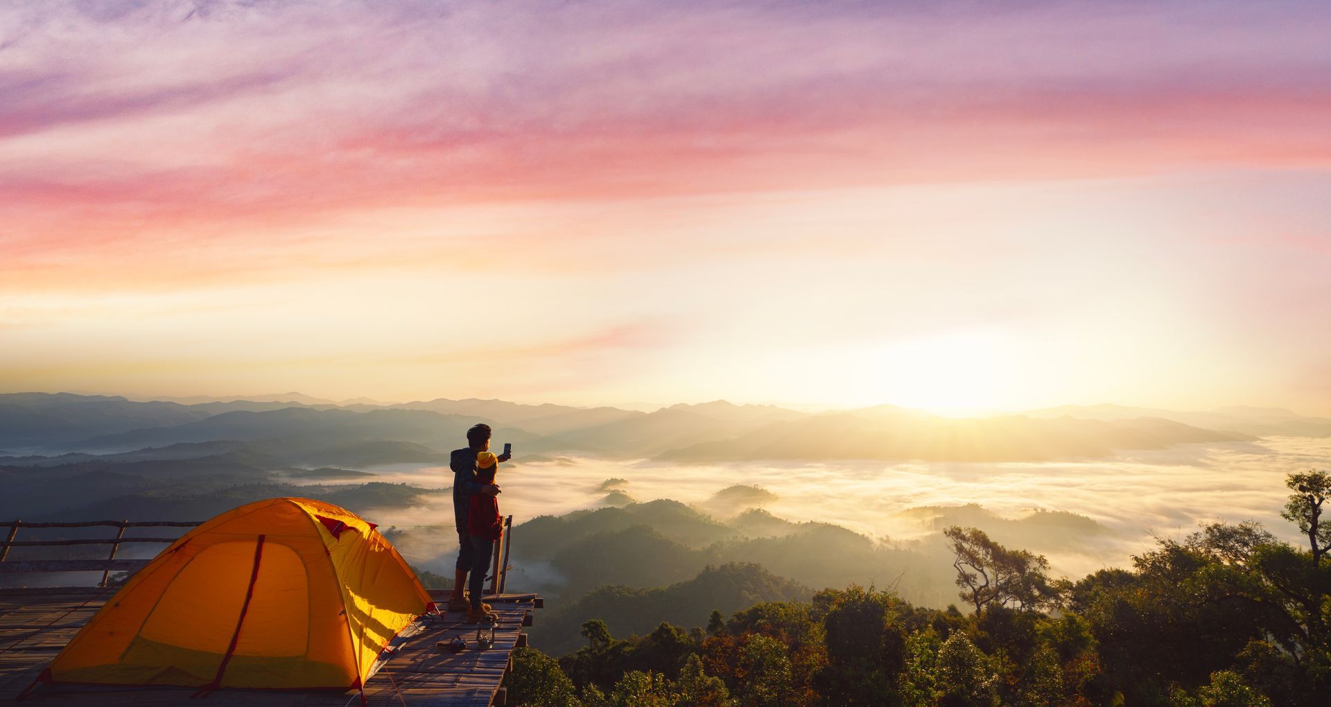 Tent on a mountain overlook at sunrise, with a person photographing a glowing sky above misty hills