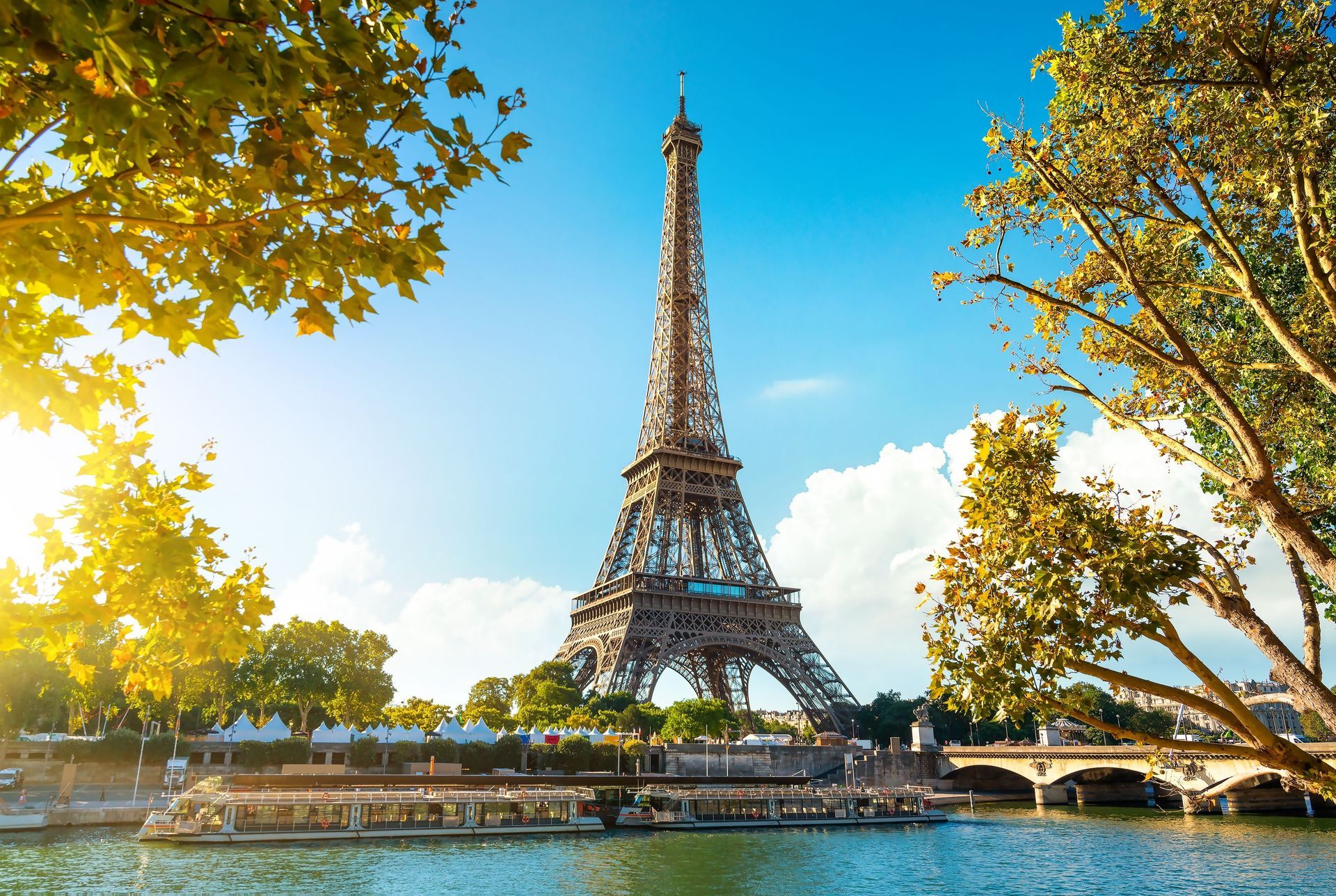 Eiffel Tower framed by autumn trees beside a river under a bright blue sky