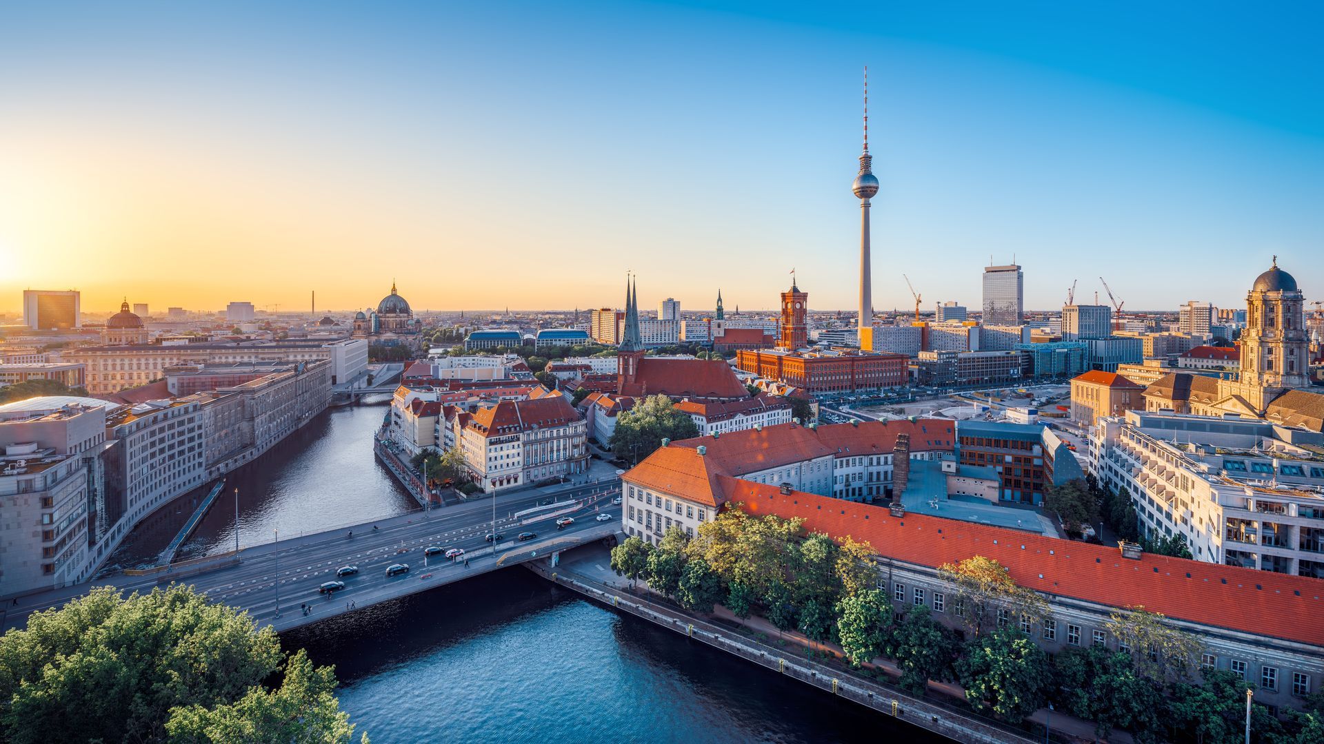 Sunset city skyline over a river and bridge, with a tall TV tower and red-roofed buildings