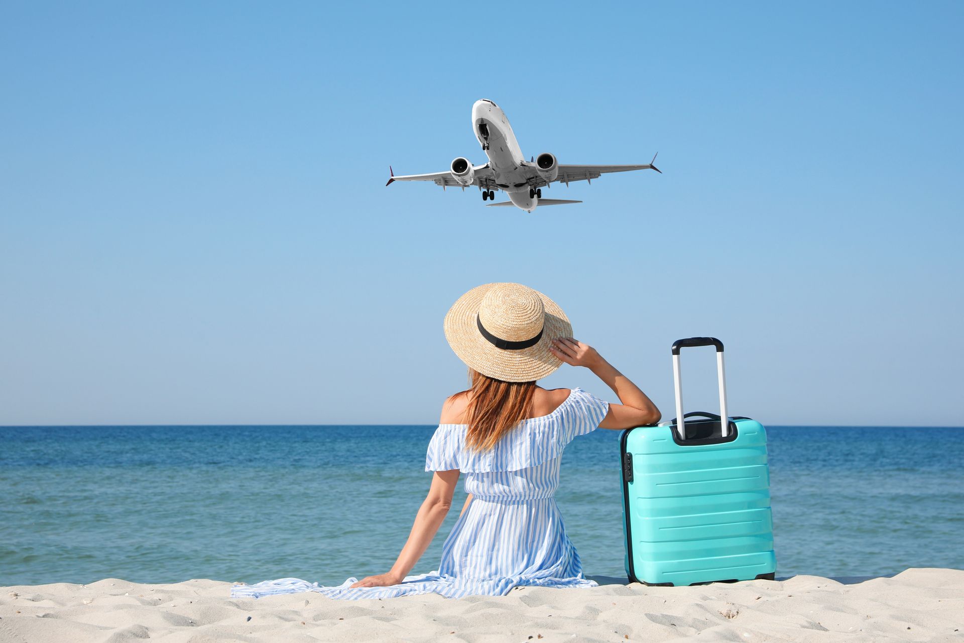 Beachgoer watching a plane take off over the ocean beside a turquoise suitcase