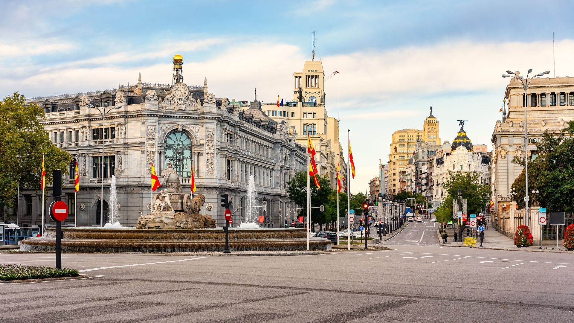 Wide city square with ornate historic buildings, a clock tower, and flags lining a mostly empty street under a bright sky