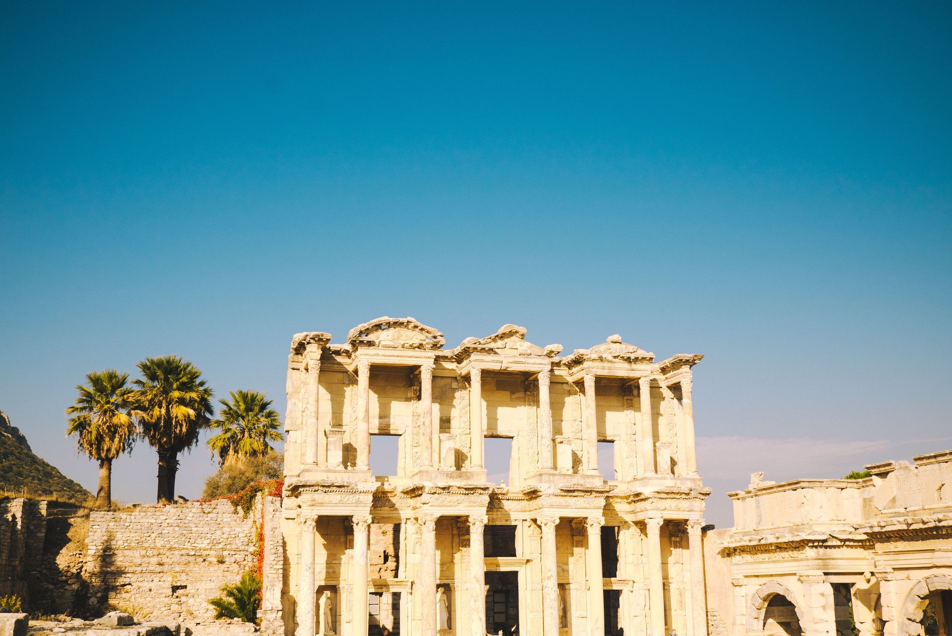 Ruins of a pale stone palace with columns under a clear blue sky, framed by palm trees
