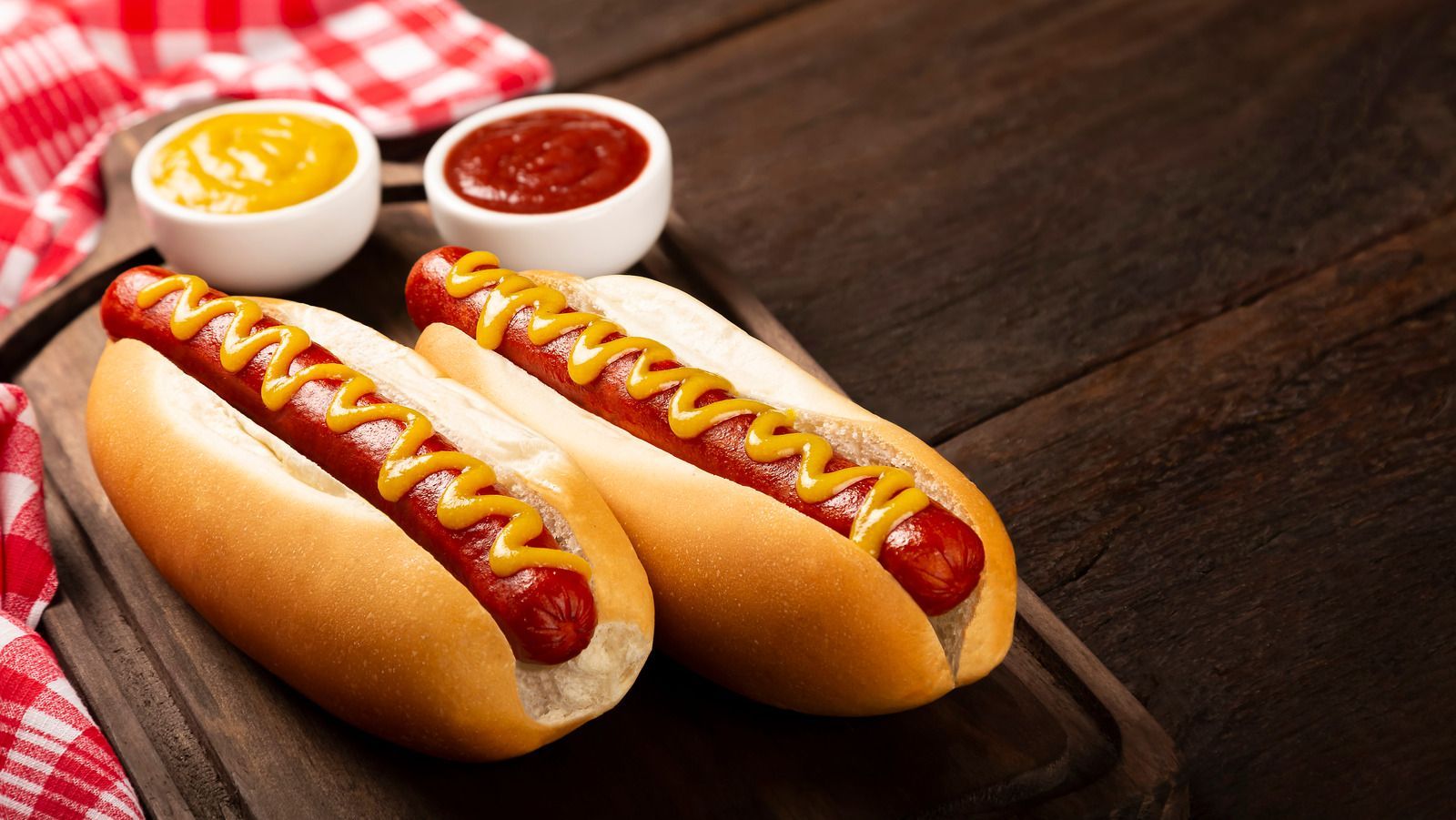 Twee hotdogs in broodjes met mosterd, ketchup en een rood geblokt tafelkleed op een houten plank.