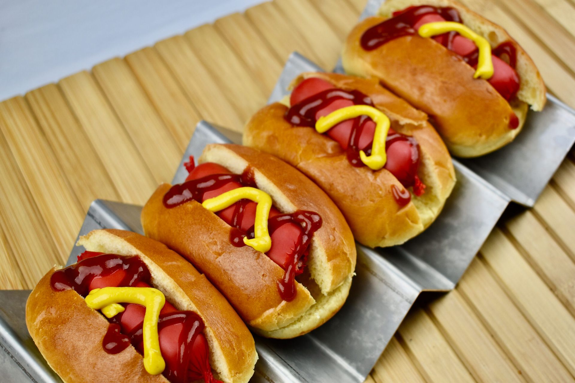 Vier hotdogs in broodjes, met ketchup en mosterd, op een metalen etagère, boven een bamboematje.