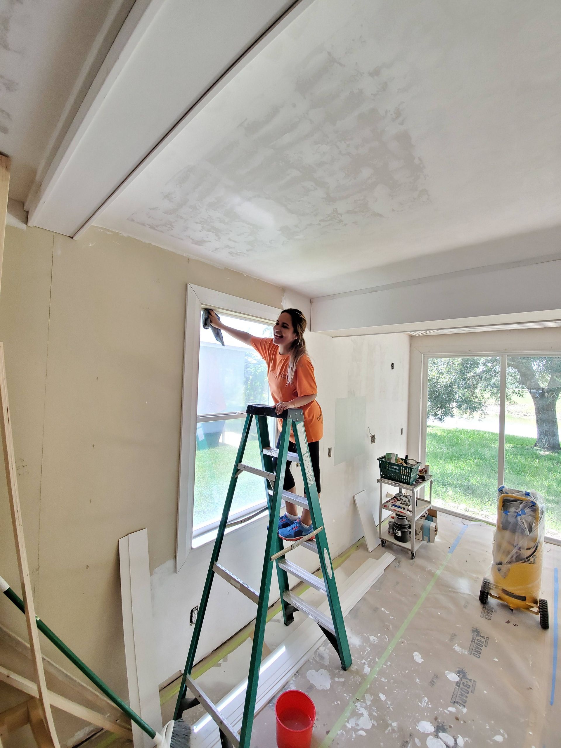 A woman is standing on a ladder in a room painting a window.