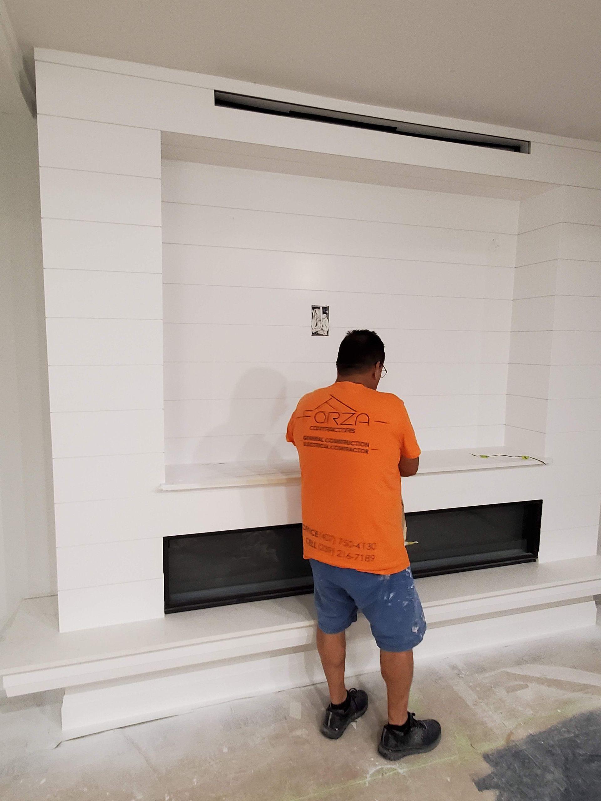 A man in an orange shirt is standing in front of a fireplace.