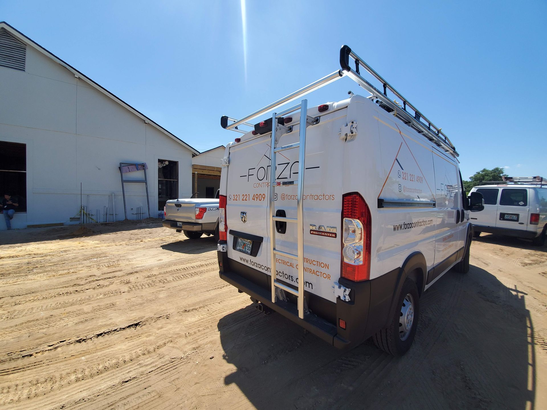 A white van with a ladder on the back is parked in a dirt lot