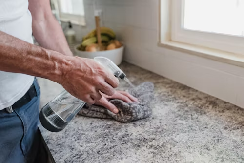 Person spraying cleaner on a countertop and wiping it with a cloth near a window.