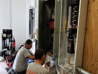 Two electricians installing electrical panel in a room with tools and supplies.
