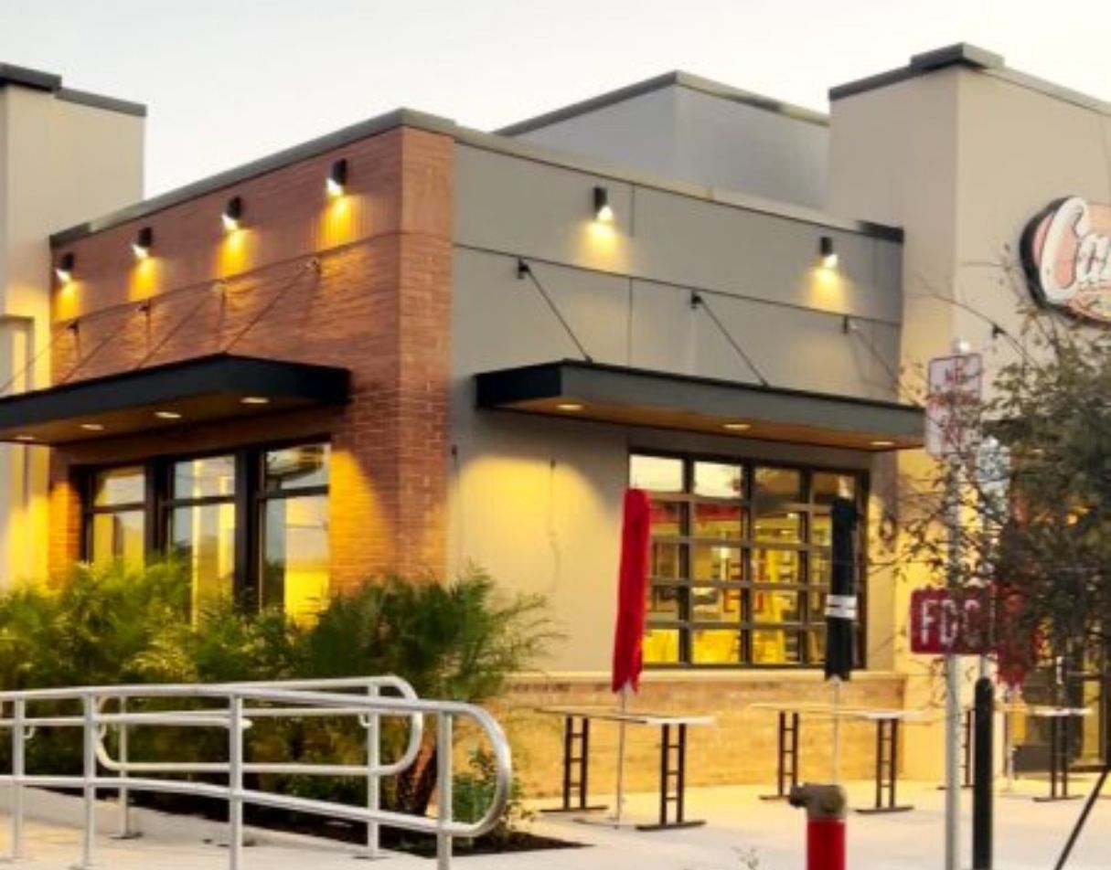 Exterior of a Cane's restaurant at dusk, featuring a brick facade, large windows, and outdoor seating with a red umbrella.