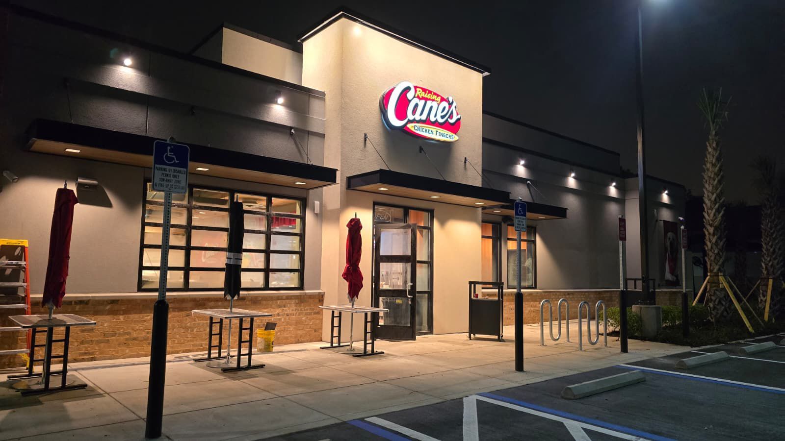Exterior of Raising Cane's restaurant at night. Illuminated sign, closed entrance, empty outdoor tables, gray facade.