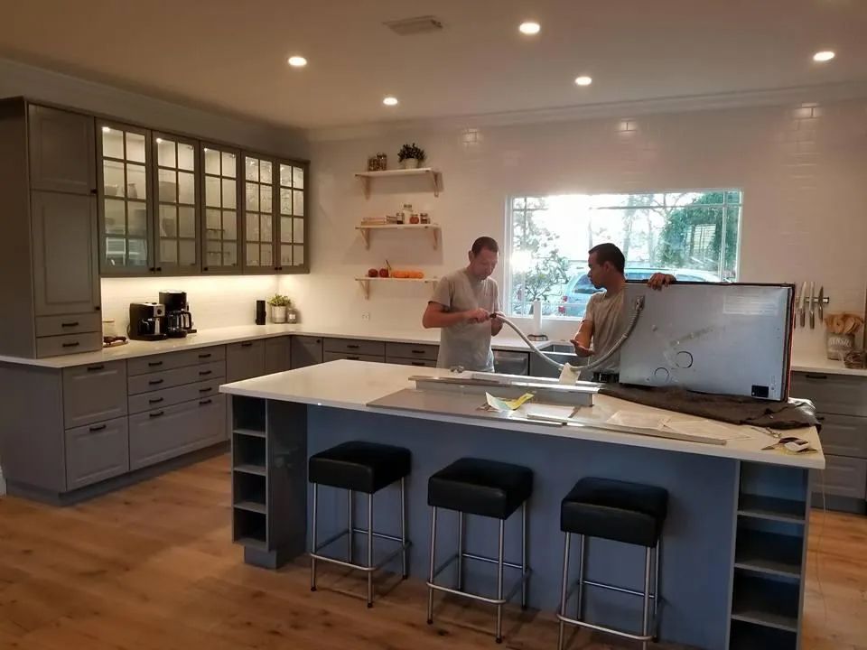 Two men work in a modern kitchen with gray cabinets, a large island, and wood floors. One man holds a white object.