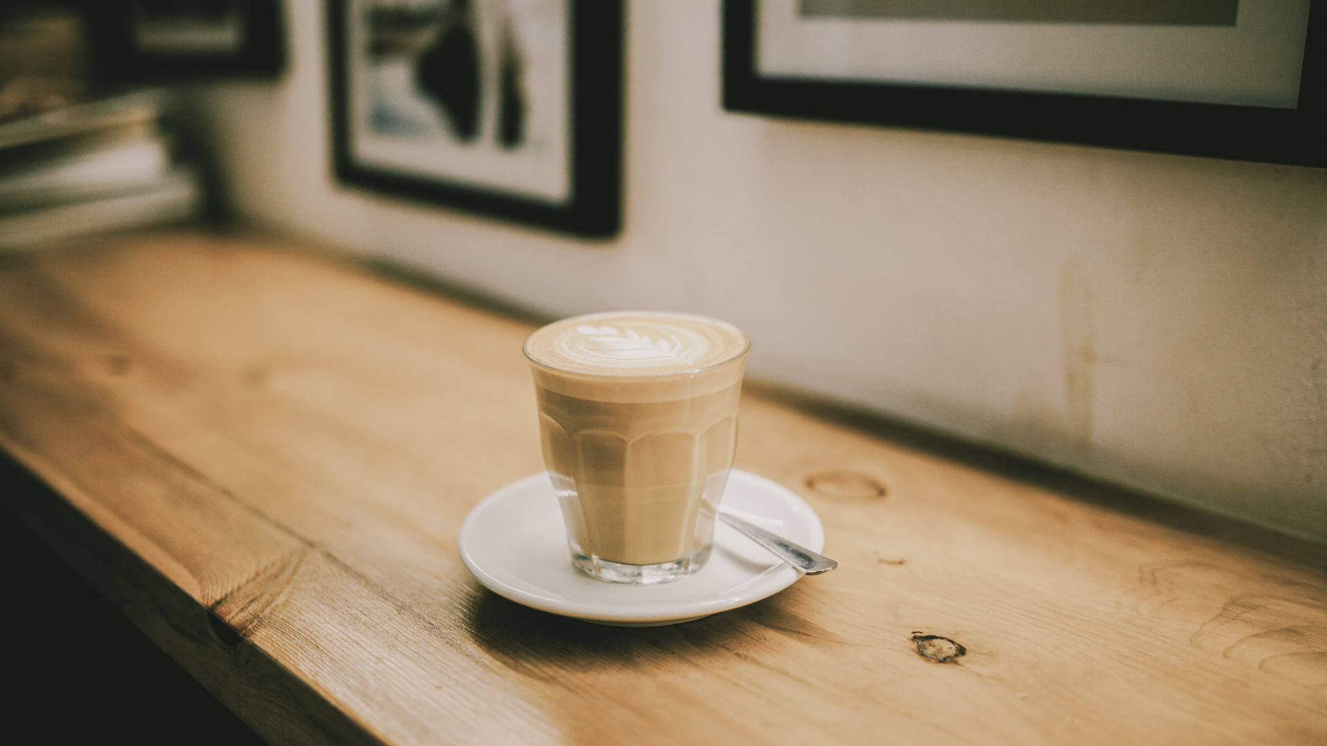 A glass of latte with foam art sits on a small white saucer on a light wood counter in a cozy cafe.