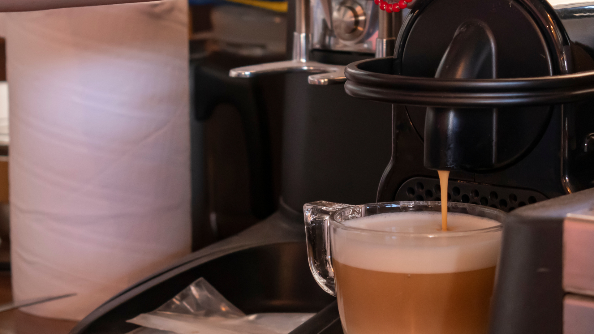 A black espresso machine dispenses a thin stream of coffee into a glass mug filled with milk foam.
