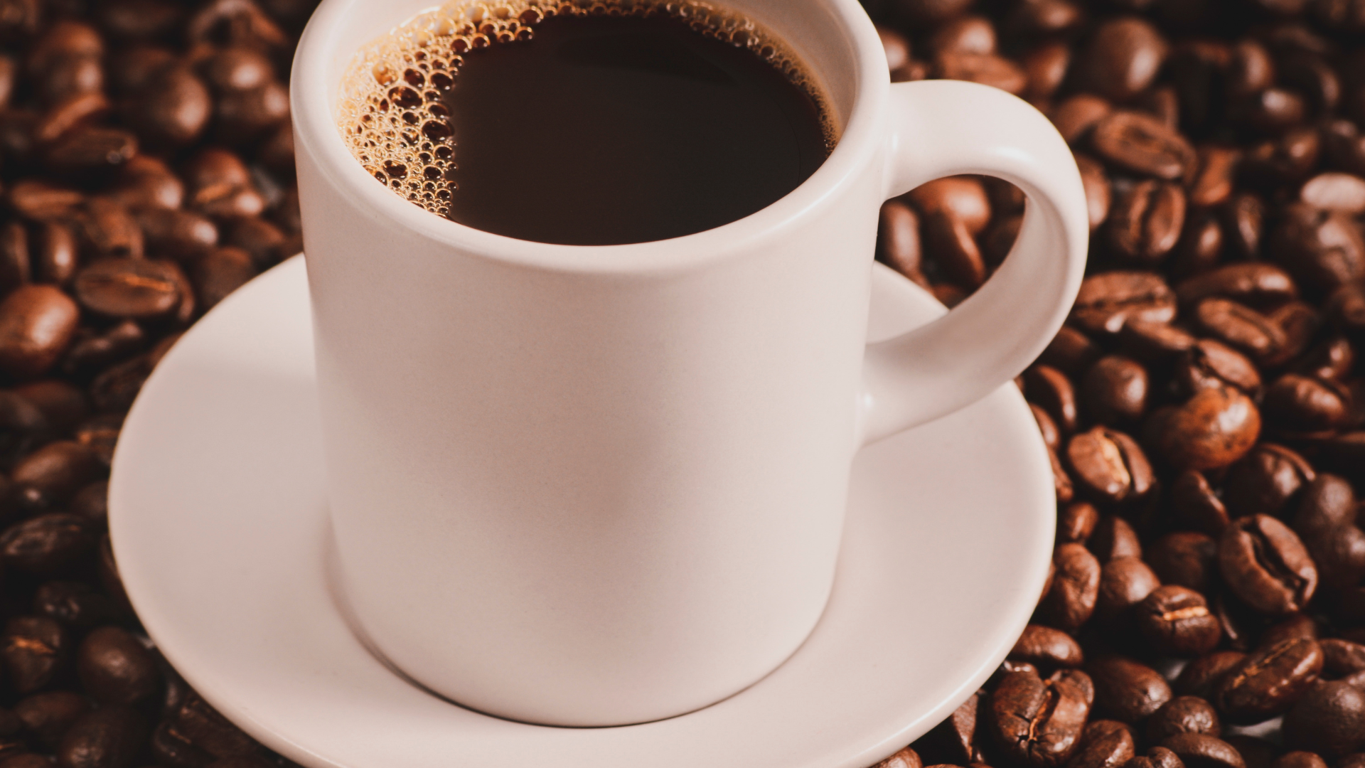 A white mug filled with black coffee sits on a matching saucer surrounded by a large pile of roasted coffee beans.
