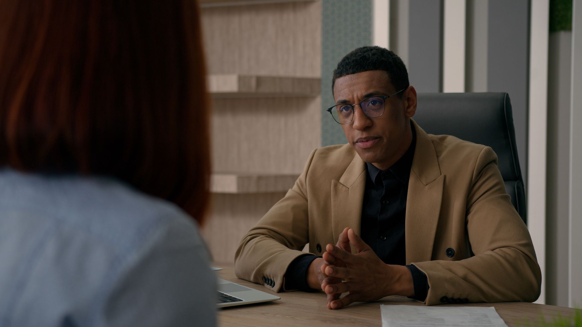 Two people in a serious office meeting across a desk, one speaking while the other listens.