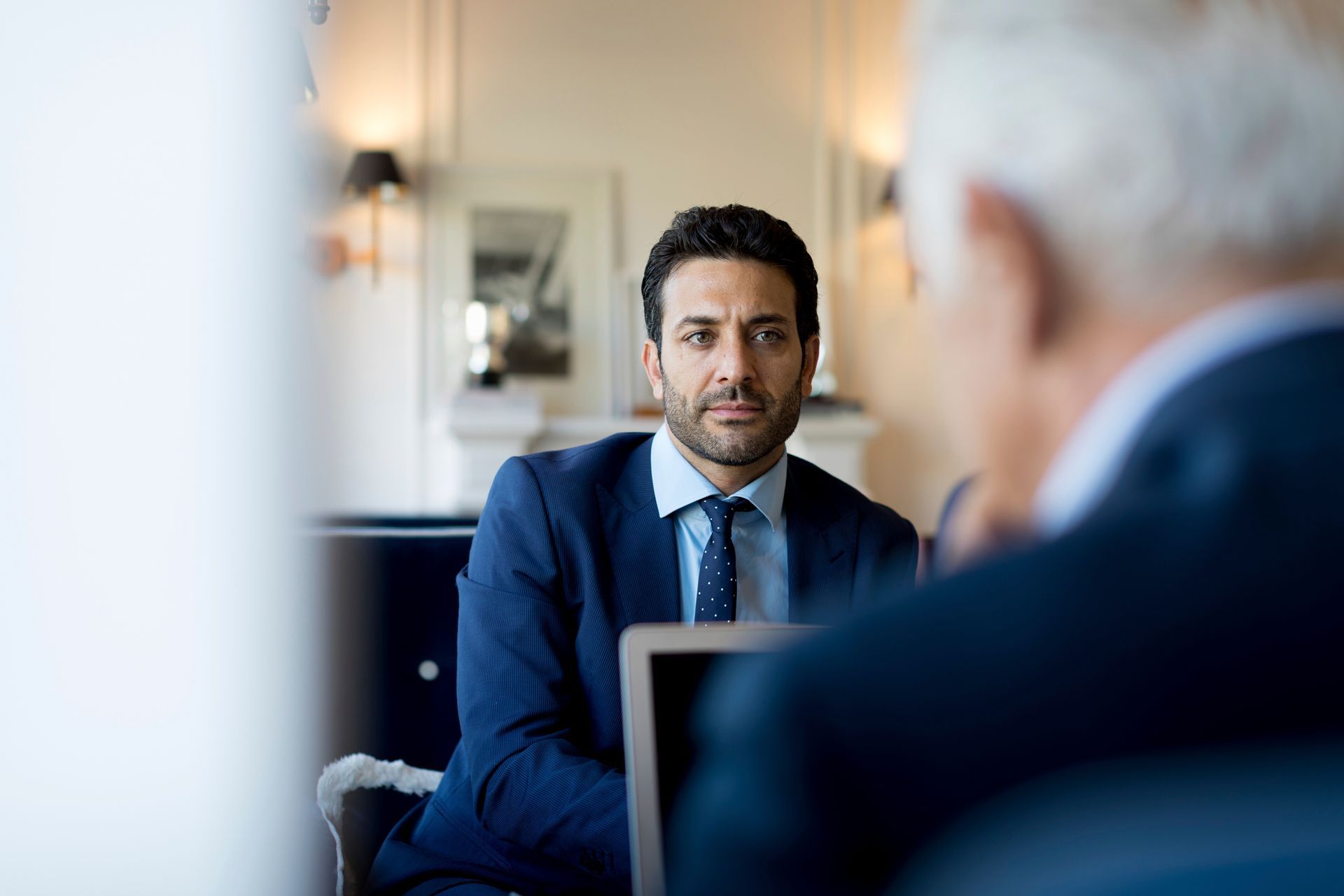 Man in a navy suit speaking in a blurred office meeting setting