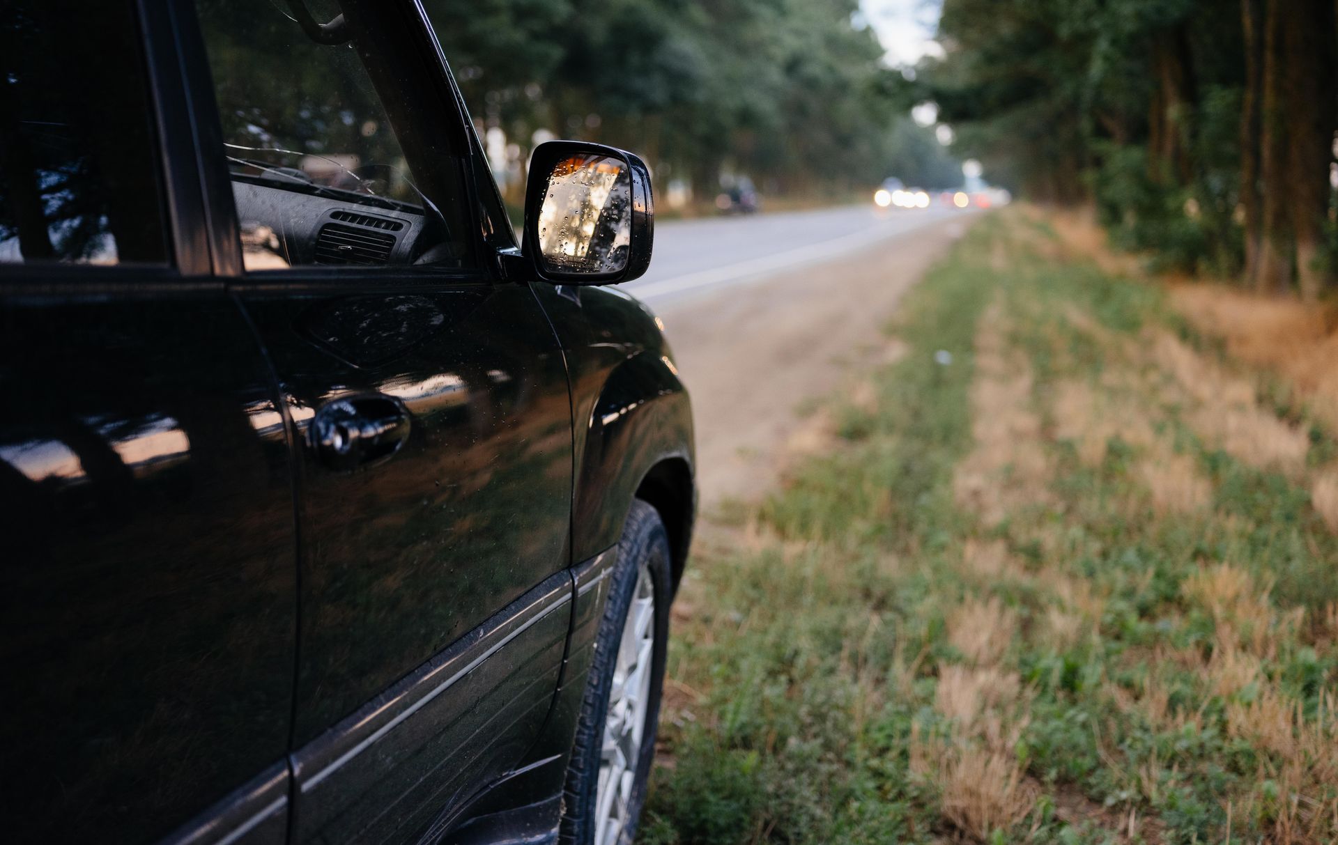 Black car parked beside a grassy roadside with trees lining the road