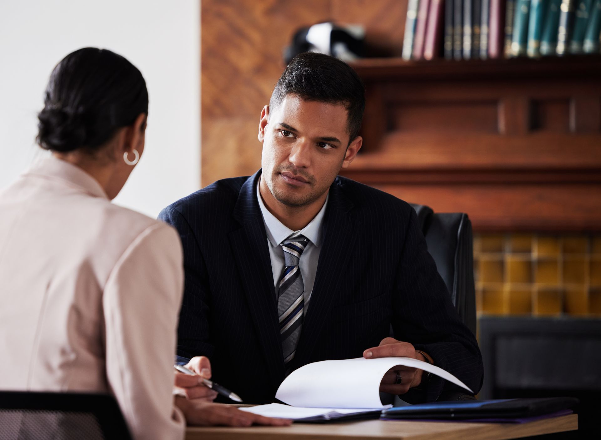 Two people in a serious discussion at a desk, with documents in a formal office setting.