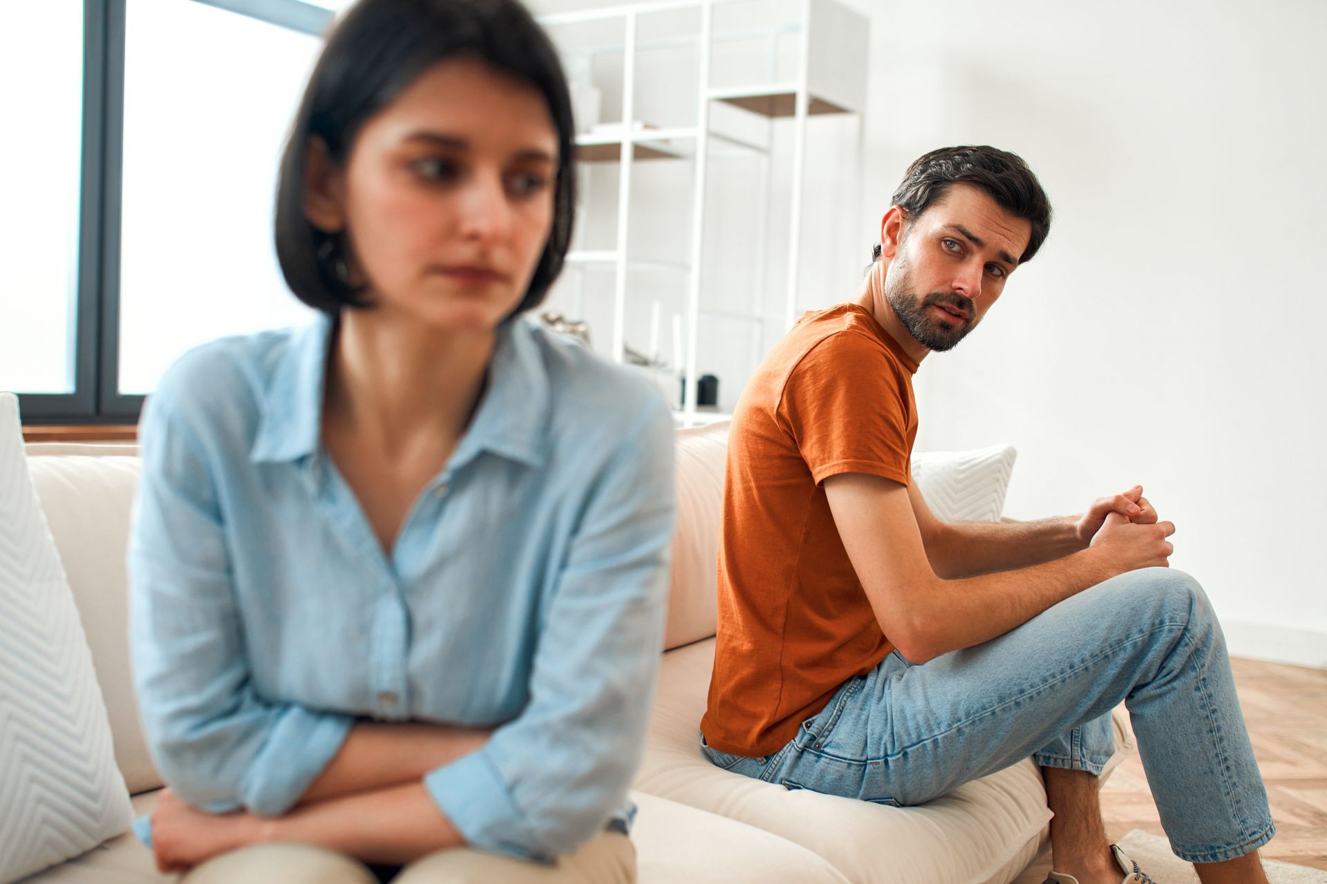 Two people sit apart on a couch, both looking away with tense expressions in a bright room.