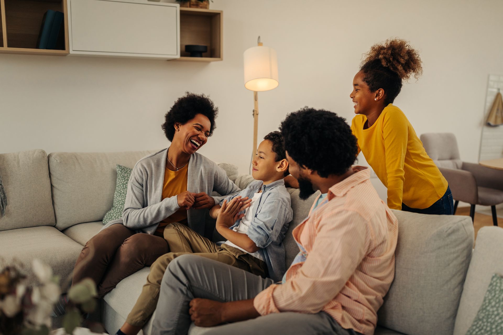 Friends chatting on a beige sofa in a cozy living room, with a floor lamp and shelves in the background.