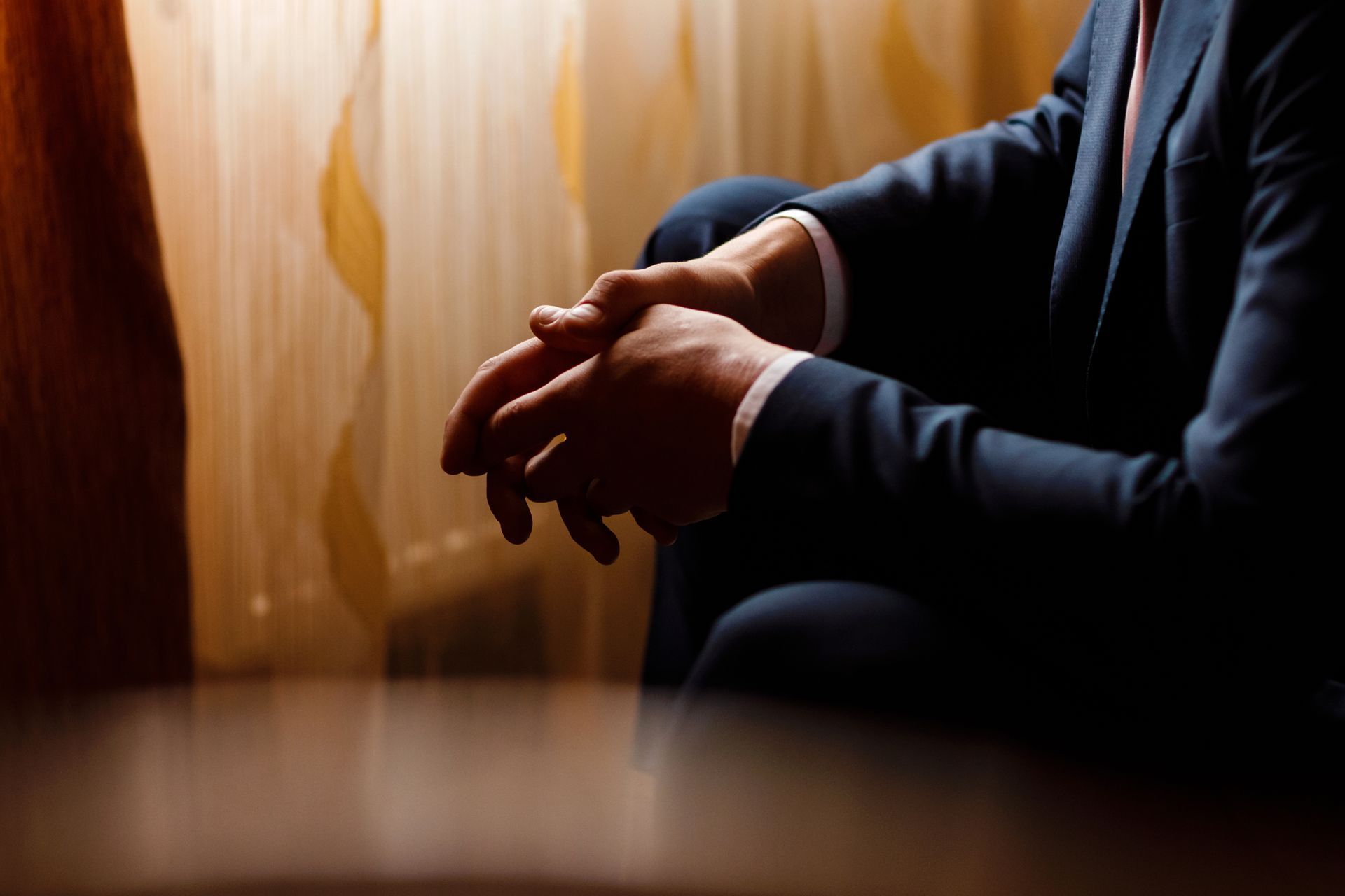 Hands clasped in a dimly lit room beside a softly lit curtain
