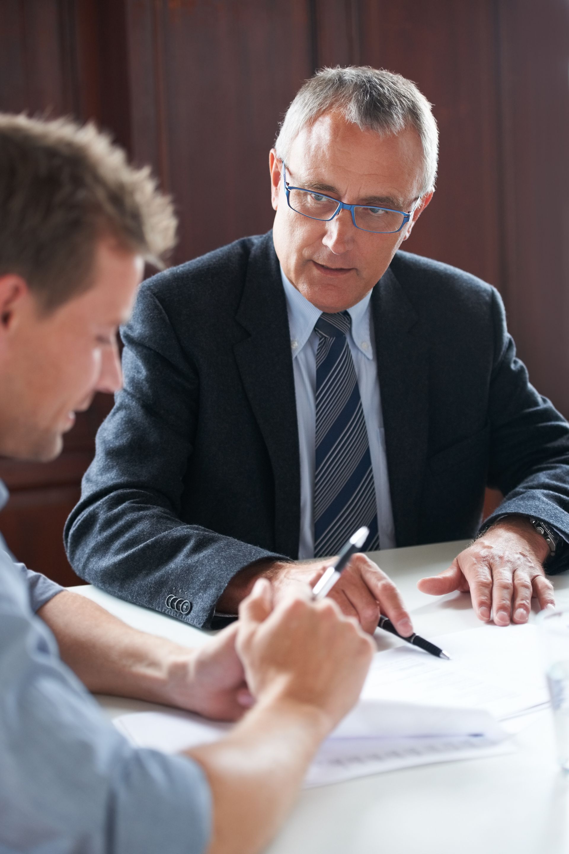 Two people in a meeting, one older man in a suit speaking at a table with papers and a pen
